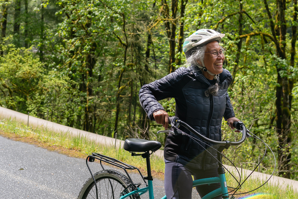 A senior woman in activewear and helmet walks her bicycle along a road lined with trees