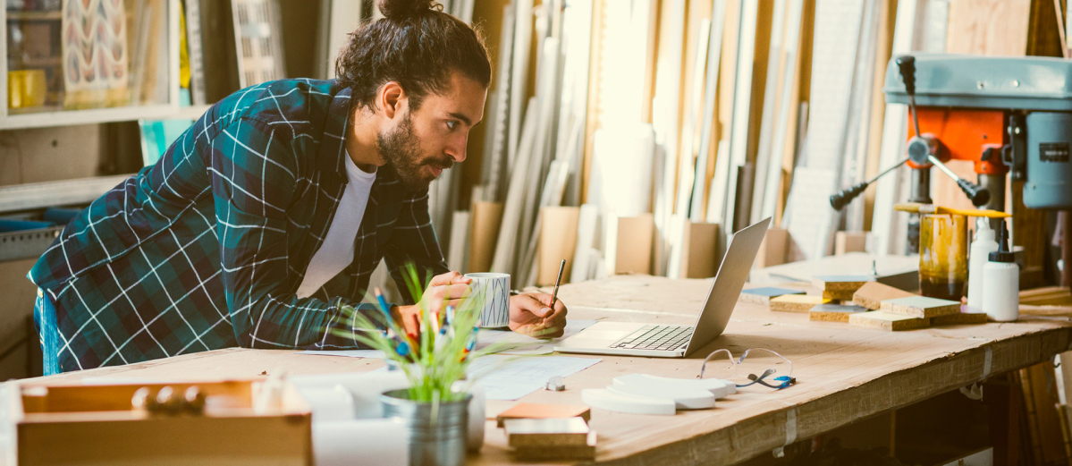 Male carpenter in workshop with coffee looking at laptop