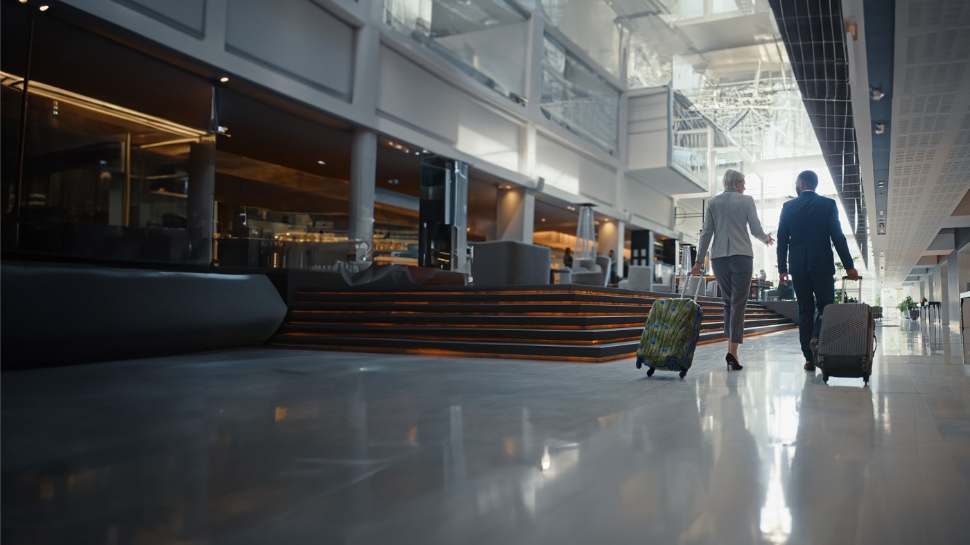 Two business professionals walking together through a hotel lobby, wheeling two small suitcases.