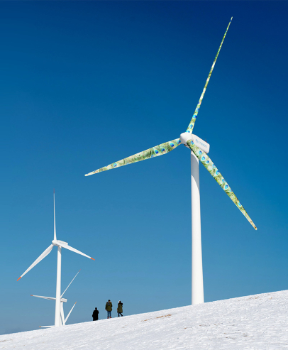 Three people walking past two wind turbines on a snowy field, one of which has a peacock feather pattern on the blades.